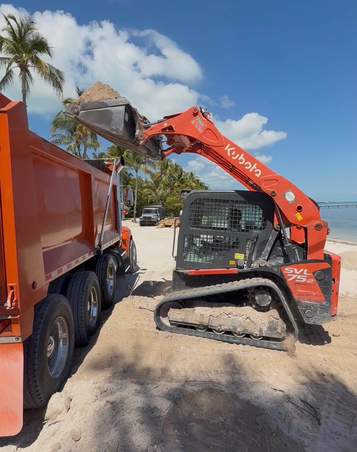Kubota SVL75-2 skid-steer loading coral rock material into a dump truck on a Florida Keys oceanfront job site — Alex Landscaping, Marathon FL.